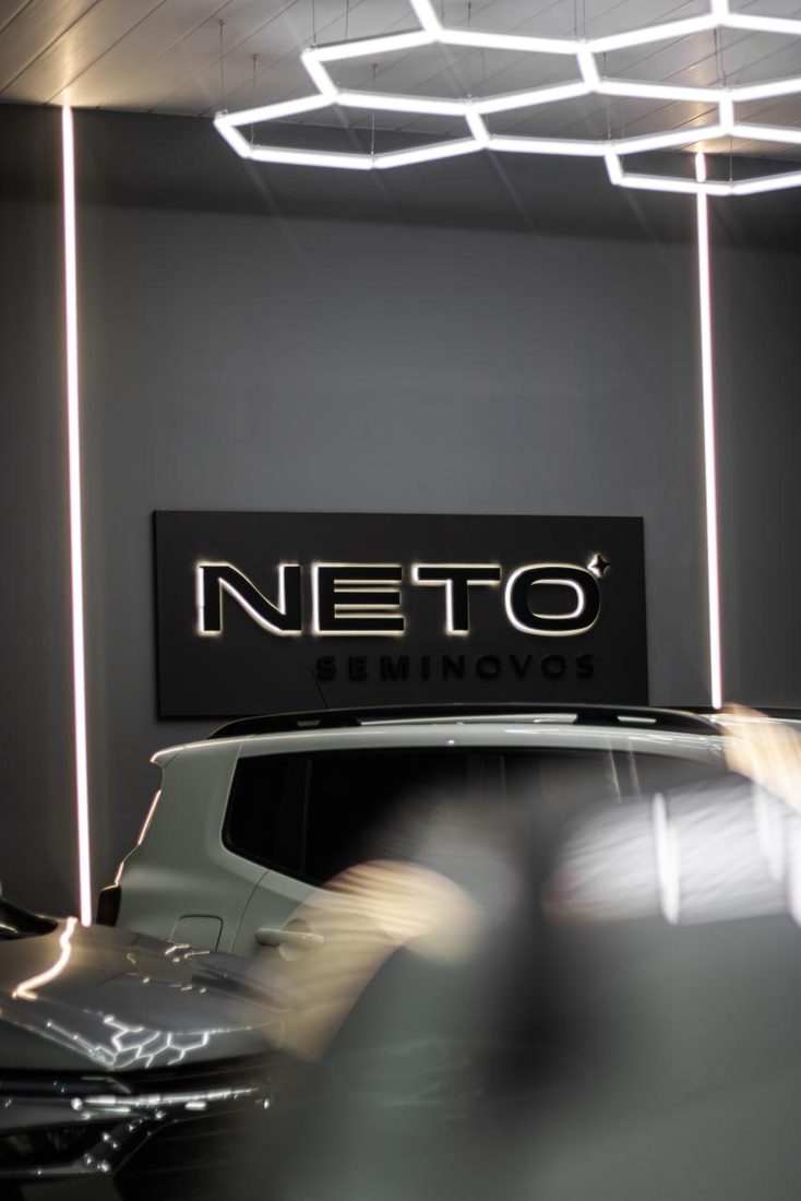 Letreiro iluminado da Neto Seminovos dentro do salão, com um veículo à frente
